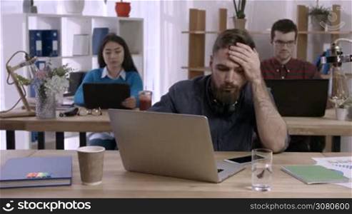 Sleepy tired freelancer with beard falling asleep at desk in front of laptop in modern office. Laughing diverse colleagues throwing crumpled paper in sleepy hipster coworker to wake him up at office.
