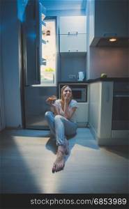 Sleepy woman sitting on floor at open fridge at late night