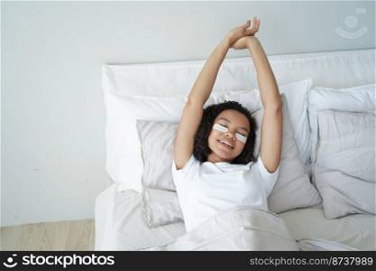 Sleepy young mixed race girl with under eye patches stretching, lying in bed. Happy biracial woman stretches after awaking in bedroom. Skincare, beauty routine, healthy lifestyle concept.. Sleepy young mixed race girl with under eye patches stretching, lying in bed. Skincare, wellness