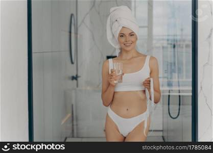 Slender happy athletic woman in white classic underwear standing in bathroom after shower routine with measuring tape in one hand and holding glass of water in other hand, healthy lifstyle concept. Slender happy athletic woman in white classic underwear standing in bathroom with measuring tape