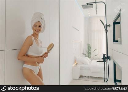 Slender happy female model posing with head wrapped in towel leaning against bathroom wall and holding wooden brush, preparing for morning shower and hygiene routine. Women beauty and body care. Slender young female model posing with head wrapped in white towel