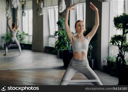 Slender redhead fitness instructor enjoys squats with pilates resistance band in gym, hands raised overhead. Workout joyfully amid mirrored walls.