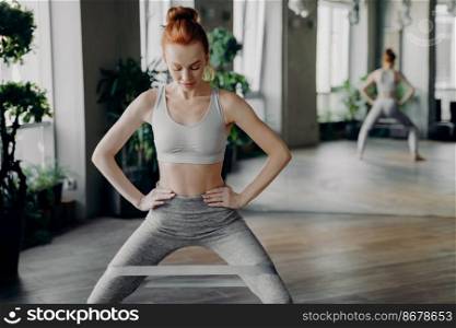 Slender sportive redhead lady performing exercises with fitness elastic band on legs, keeping hands on waist, looking down at toes in gym with large mirror behind. Healthy lifestyle and sport indoor. Slender sportive redhead lady performing sit ups with fitness elastic band