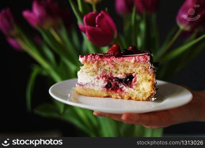 slice of berry cake on a white plate near beautiful tulips flowers. young woman holding a plate with a slice of cake in her hand.. slice of berry cake on a white plate near beautiful tulips flowers. young woman holding a plate with a slice of cake in her hand