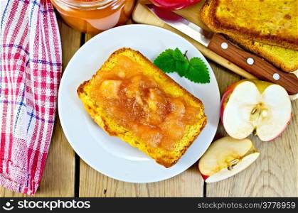Slice of bread with apple jam and mint on a plate, slices of apple, a jar of jam, napkin, knife on background wooden plank