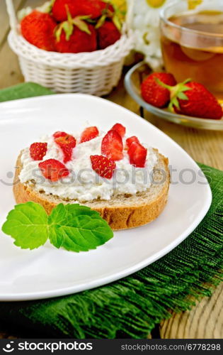 Slice of french bread with curd cream, strawberry and mint on a plate and a napkin, basket with berries, glass cup with tea on a wooden board