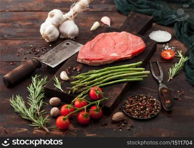 Slice of fresh raw barbeque braising beef steak on chopping board with asparagus and garlic with tomatoes and salt with pepper on wooden background with hatchet.