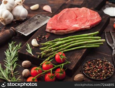 Slice of fresh raw barbeque braising beef steak on chopping board with asparagus and garlic with tomatoes and salt with pepper on wooden background with hatchet.