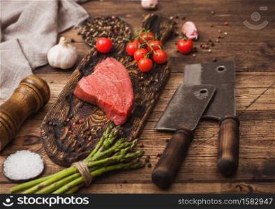 Slice of Raw Beef sirlion steak on wooden chopping board with tomatoes,garlic and asparagus tips and meat hatchets on wooden kitchen table background.