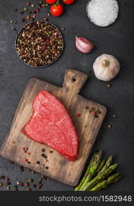 Slice of Raw Beef sirlion steak on wooden chopping board with tomatoes,garlic and asparagus tips on dark kitchen table background.