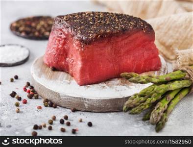 Slice of Raw Beef Topside Joint with Salt and Pepper on round chopping board with asparagus tips and garlic on light kitchen table background.