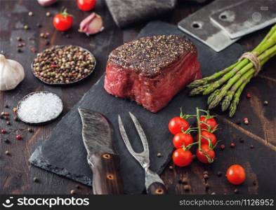 Slice of Raw Beef Topside Joint with Salt and Pepper on stone chopping board with fork and knife, garlic and asparagus tips and tomatoes on wood kitchen table.