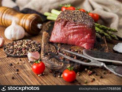 Slice of Raw Beef Topside Joint with Salt and Pepper on wooden chopping board with fork and knife, asparagus tips and kitchen towel on wood kitchen table.