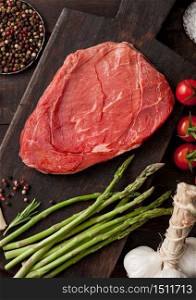 Slice of raw fresh beef braising steak on chopping board with garlic, asparagus and tomatoes with salt and pepper on wooden table background. Macro