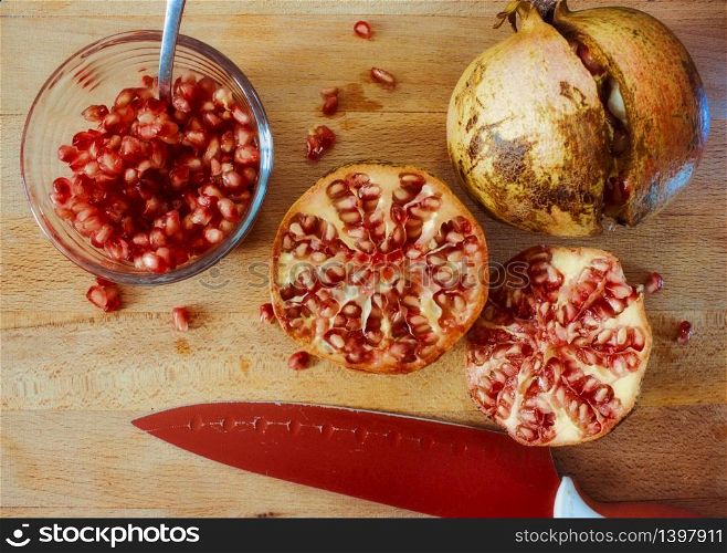 Sliced and intact pomegranates on a wooden chopping board with a glass bowl of seeds for juicing and knife