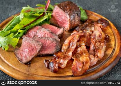 Sliced beef steak with fried rashers of bacon and leafs of fresh arugula on the wooden board