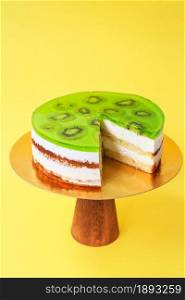 Sliced Birthday cake decorated with green jelly and kiwi on top on the wooden cake stand. Beautiful sponge cake with whipped cream. Yellow background. Copy space. Food photography for recipe.