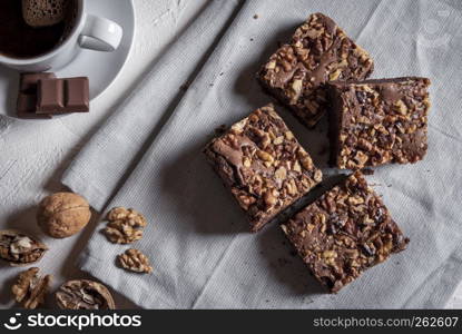 Sliced brownie cake decorated with walnuts on a towel. Tabletop view with a cup of coffee, chocolate and walnuts, near the delicious cake