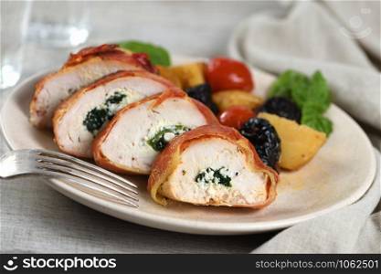 Sliced chicken breast stuffed with goat cheese with spinach, wrapped in prosciutto, with a side dish of baked potatoes, tomato and dried prunes