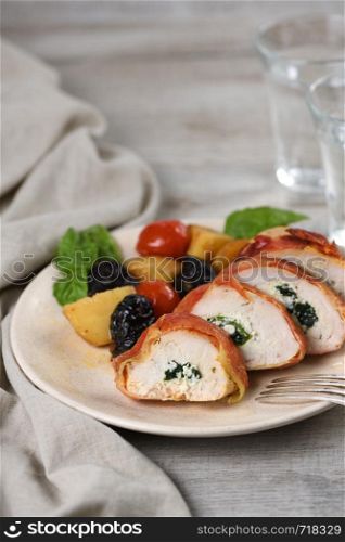 Sliced chicken breast stuffed with goat cheese with spinach, wrapped in prosciutto, with a side dish of baked potatoes, tomato and dried prunes
