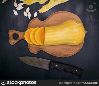 sliced fresh peeled pumpkin on brown cutting board next to a knife, black background, top view