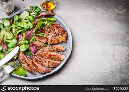 Sliced grilled beef steak with green leaves salad on rustic plate with cutlery. Medium rare barbecue steak and healthy salad on gray stone background, top view, place for text