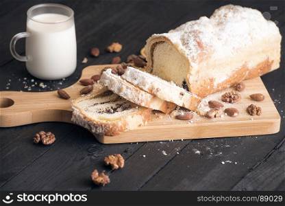 Sliced homemade sponge cake filled with poppy seeds, walnuts and almonds, displayed on a wooden trencher, with a glass of milk near, on a rustic wooden table