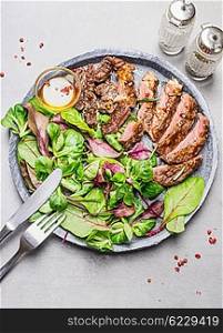 Sliced medium rare grilled barbecue steak and green salad on stone plate with cutlery, top view