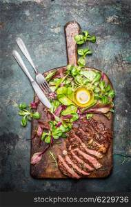 Sliced medium rare grilled beef barbecue steak served with fresh green salad and cutlery on rustic cutting board , top view. Meat food