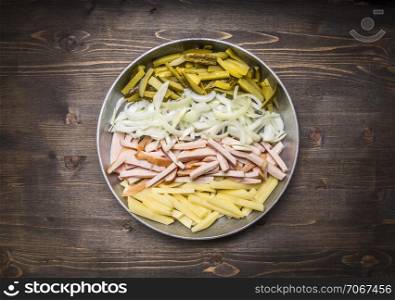 sliced onions, ham, pickles and potatoes in a frying pan on wooden rustic background top view close up