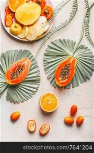 Sliced papaya with seeds on big, green leaves, various halves of orange and yellow fruits on light concrete background. Top view