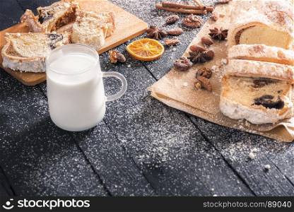 Sliced pound cake, filled with poppy seeds and walnuts, surrounded by roasted nuts and spices, covered in powder sugar, and a cup of milk on a wooden table.