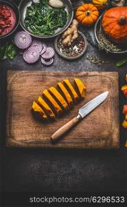 Sliced pumpkin on cutting board with knife and various vegetables and seasoning ingredients for tasty seasonal dish cooking, rustic , top view. Healthy and clean eating with organic local vegetables.