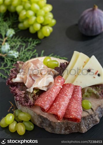 Sliced slices of salami, bacon, cheese located on a sheet of salad on a wooden board. Black stone background. Grapes and figs. Close-up.