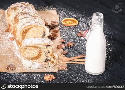 Sliced sponge cake, on a baking paper, covered in powder sugar, filled with poppy seeds and nuts, and a bottle of fresh milk, on a black wooden table.