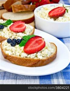 Slices of bread with curd cream, blueberries and strawberries, mint, a bowl of cottage cheese, bread on a board on a background of the blue checkered tablecloth