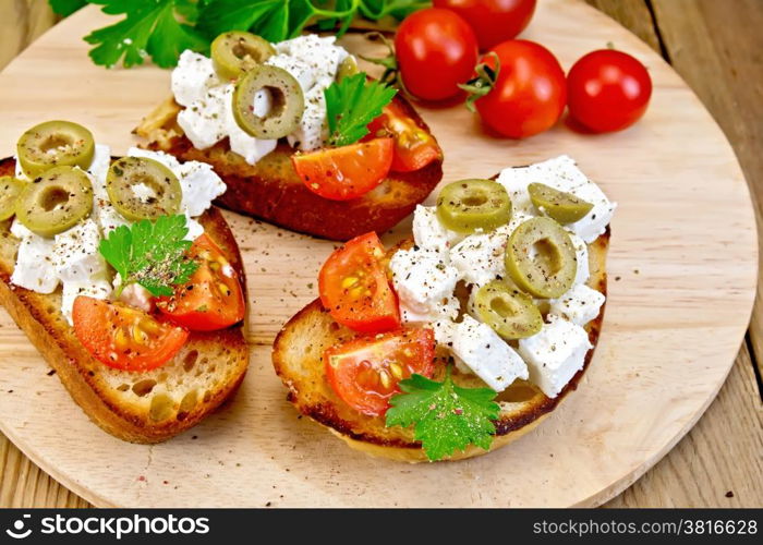 Slices of bread with feta, tomato and olives on a round board, parsley and tomatoes on a wooden table