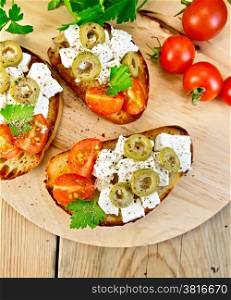 Slices of bread with feta, tomato and olives on a round board, parsley and tomatoes on wooden table background