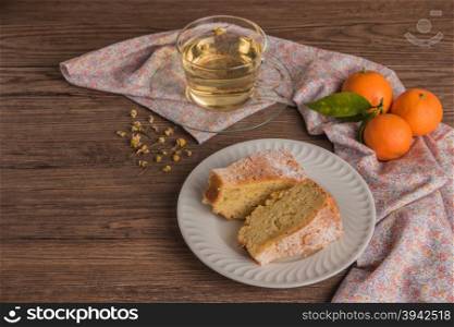 Slices of clementine cake with powdered sugar topping and cup of chamomile tea. Cake on a plate with fresh clementines on wooden board.