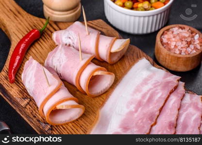 Slices of delicious raw or salted bacon with spices, salt, vegetables and herbs on a wooden cutting board against a dark concrete background. Slices of delicious raw or salted bacon with spices, salt, vegetables and herbs on a wooden cutting board