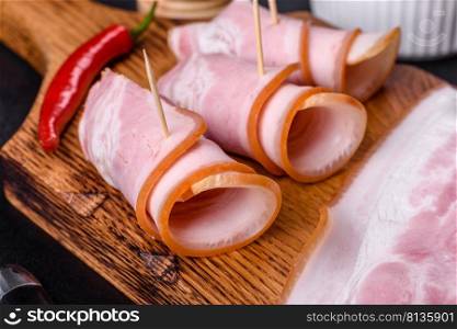 Slices of delicious raw or salted bacon with spices, salt, vegetables and herbs on a wooden cutting board against a dark concrete background. Slices of delicious raw or salted bacon with spices, salt, vegetables and herbs on a wooden cutting board