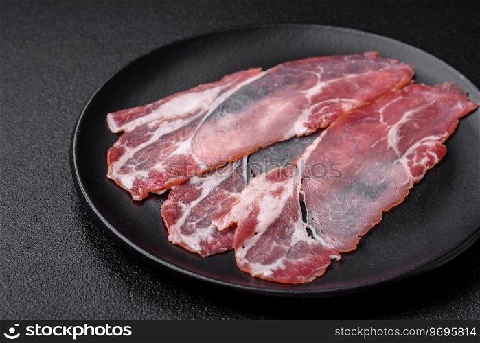 Slices of delicious smoked jamon or prosciutto with salt, spices and herbs on a dark concrete background