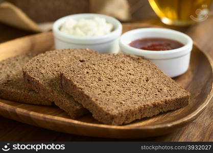 Slices of pumpernickel dark rye bread on wooden plate with jam and cream cheese, cup of tea in the back, photographed with natural light (Selective Focus, Focus diagonally in the middle of the slice)