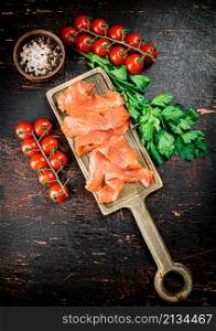 Slices of salted salmon with greens and tomatoes on a cutting board. Against a dark background. High quality photo. Slices of salted salmon with greens and tomatoes on a cutting board.