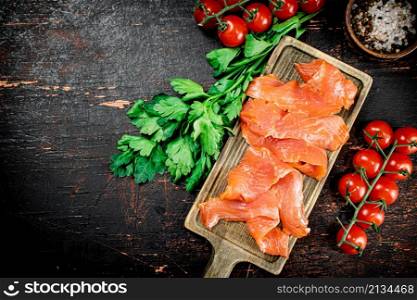 Slices of salted salmon with greens and tomatoes on a cutting board. Against a dark background. High quality photo. Slices of salted salmon with greens and tomatoes on a cutting board.