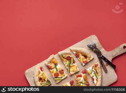 Slices of spring pizza with cherry tomatoes and zucchini, on rustic wooden platter, on a red background. Flat lay of tasty vegetables pizza primavera.