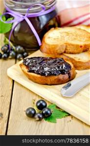 Slices of toasted bread, a glass jar with black currant jam, knife on background wooden board