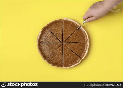Slicing a pie concept. Woman hand cutting in slices a pumpkin pie on a yellow background. Top view of delicious traditional Thanksgiving dessert.