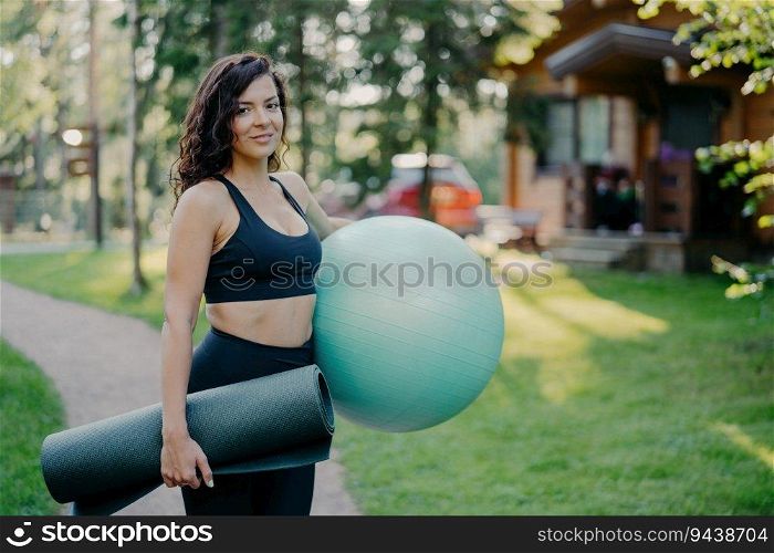 Slim active woman holds fitness ball and karemat, ready for outdoor exercises. Morning workout amid blurred forest backdrop and green grass.
