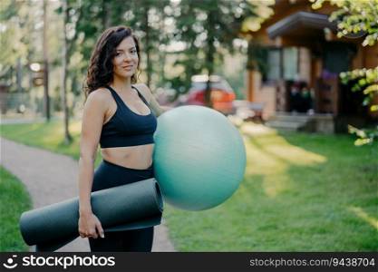 Slim active woman holds fitness ball and karemat, ready for outdoor exercises. Morning workout amid blurred forest backdrop and green grass.